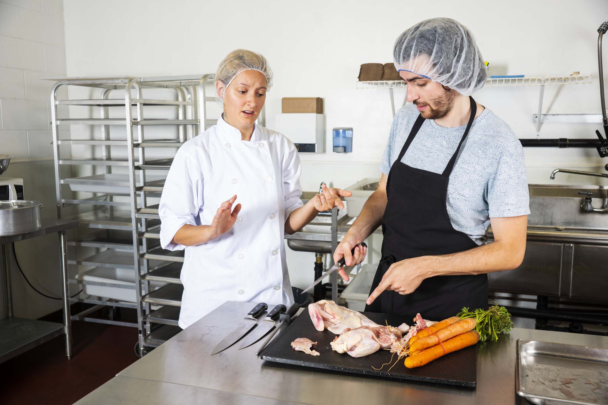 Food contamination. A food service worker cutting raw chicken on the same board beside raw vegetables. His supervisor is explaining that this is a serious safety violation. Food safety concept. Raw meat and vegetables must be separated during all phases of food storage and preparation. Food contamination class